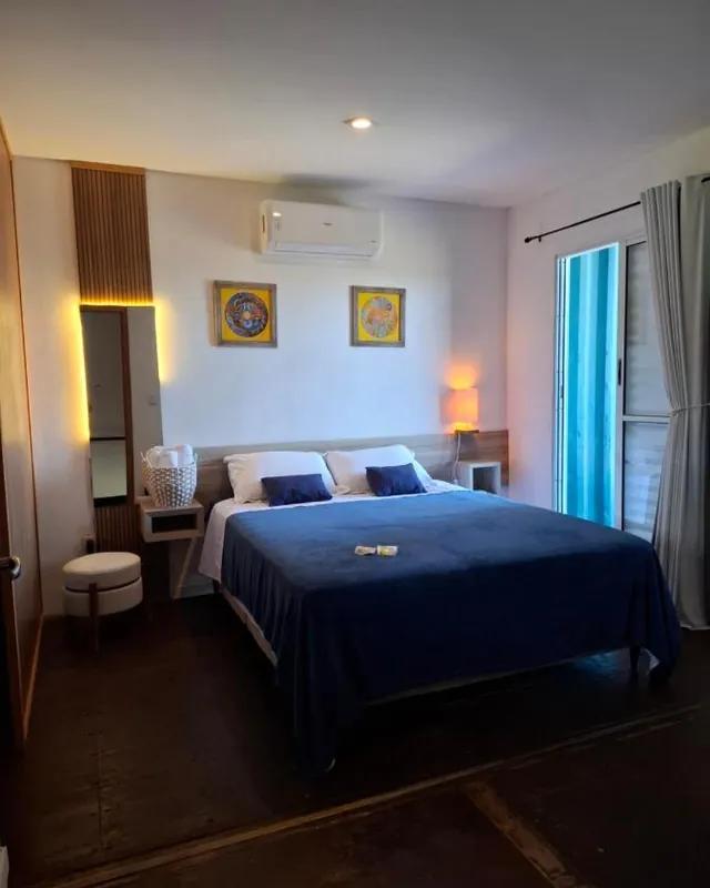 A suíte da Casa Container, com cama queen de colcha azul, abajures acesos e ar-condicionado